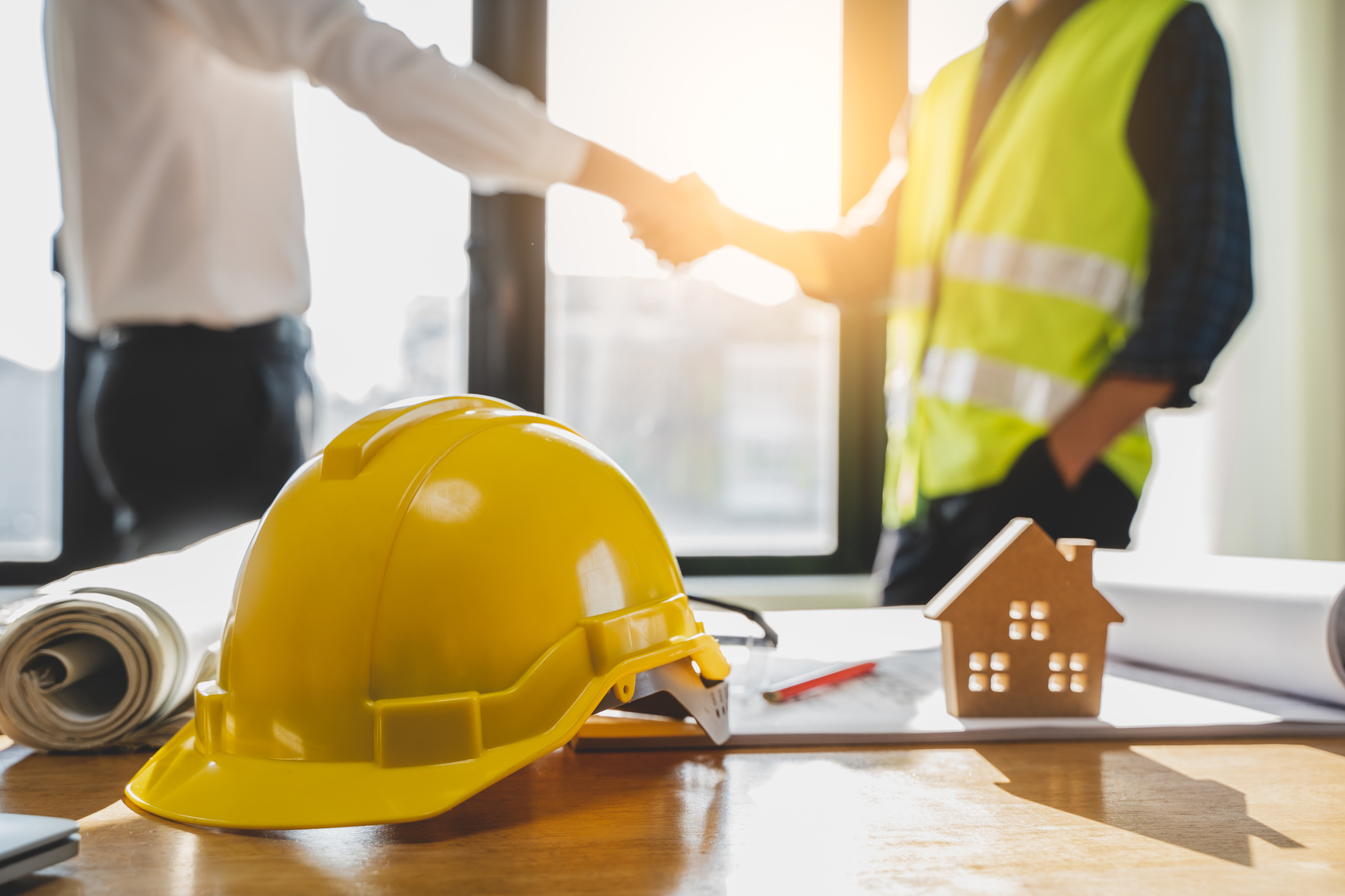 A client in business attire and a contractor in a safety vest shake hands over blueprints and a hard hat